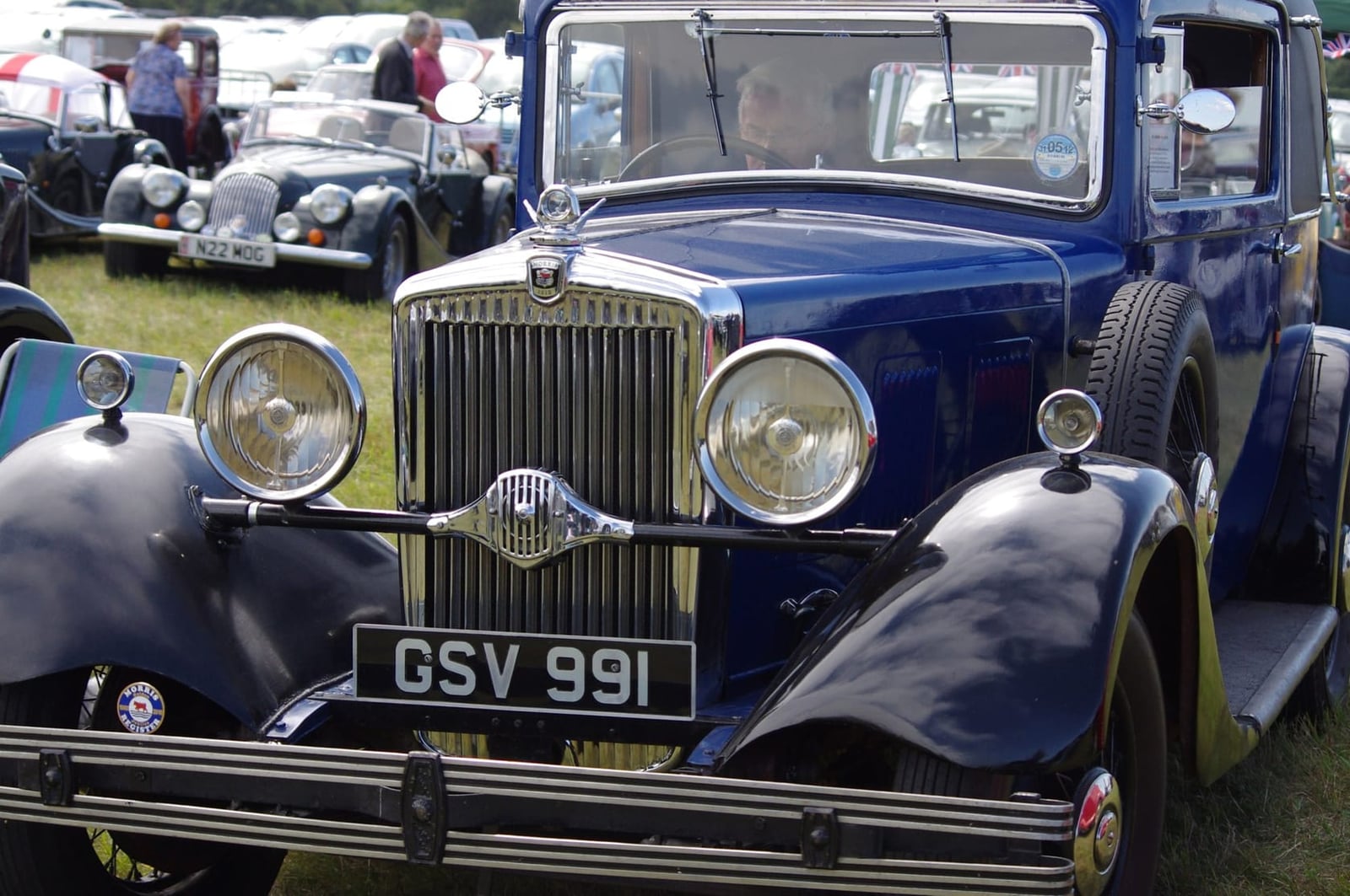 A blue 1931 Morris Major Six with chrome radiator and headlamps on grass at a vintage rally, UK registration GSV 991