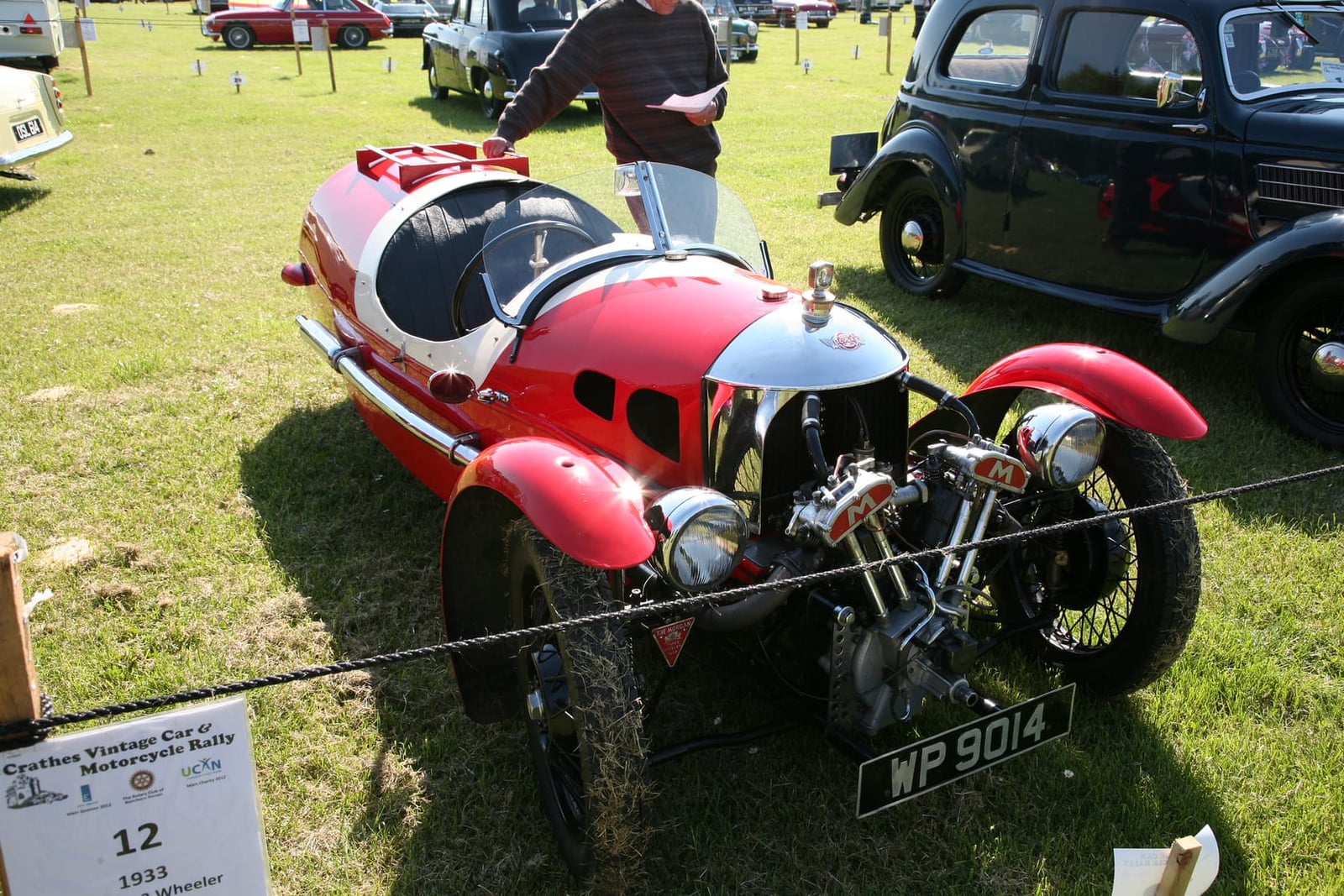 A red 1933 Morgan three-wheeler with an exposed V-twin engine on grass at a vintage car and motorcycle rally, UK registration WP 9014