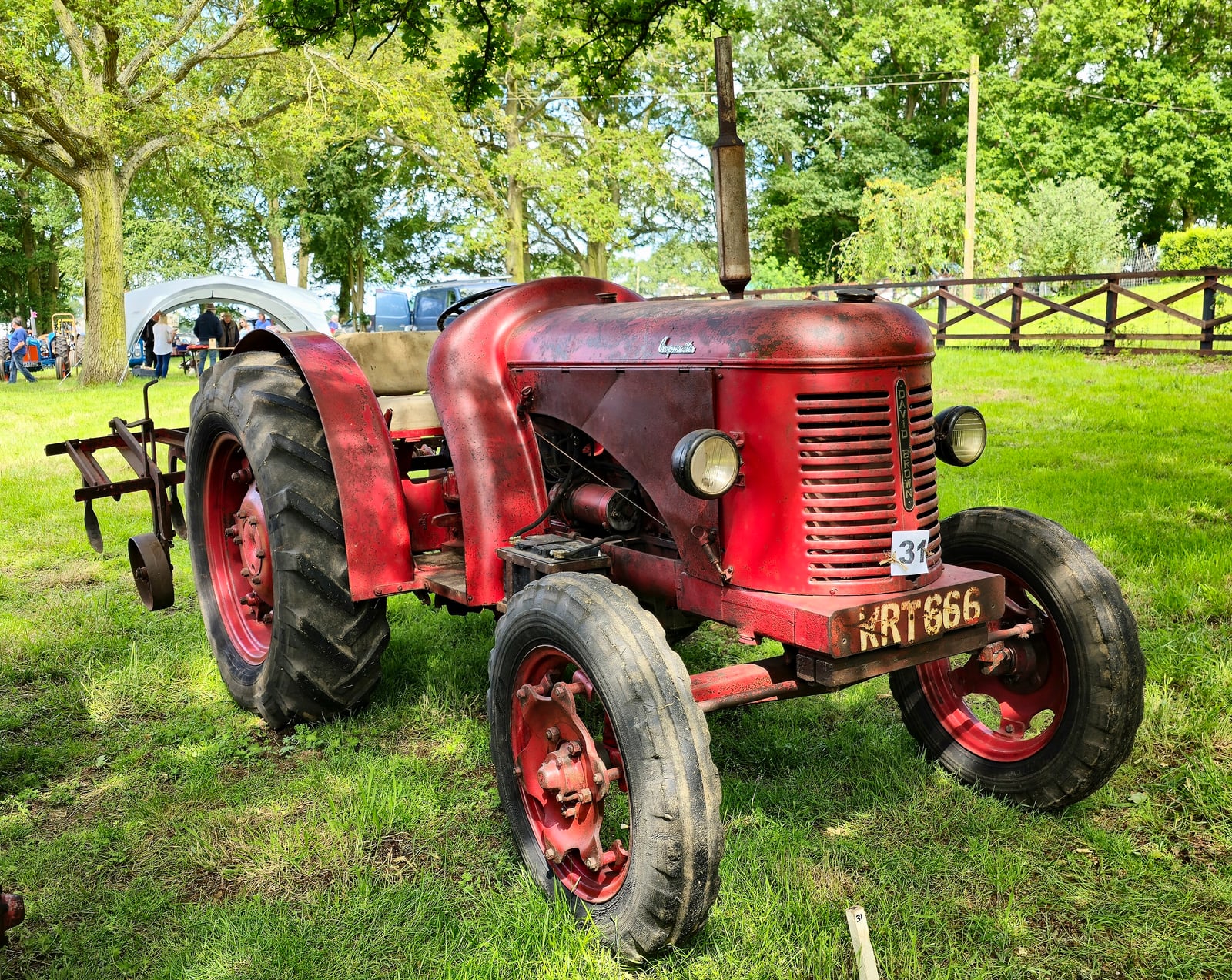 A faded works-red David Brown Cropmaster with a plough, registration RRT 666, on grass at a show