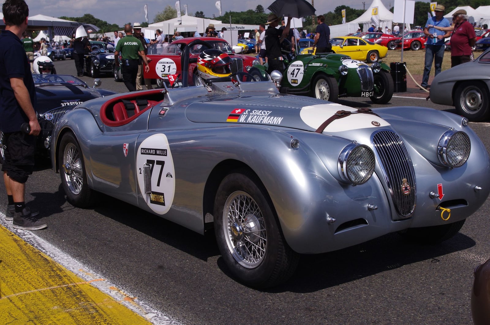 A silver Jaguar XK120 racing roadster, number 77, in a race paddock