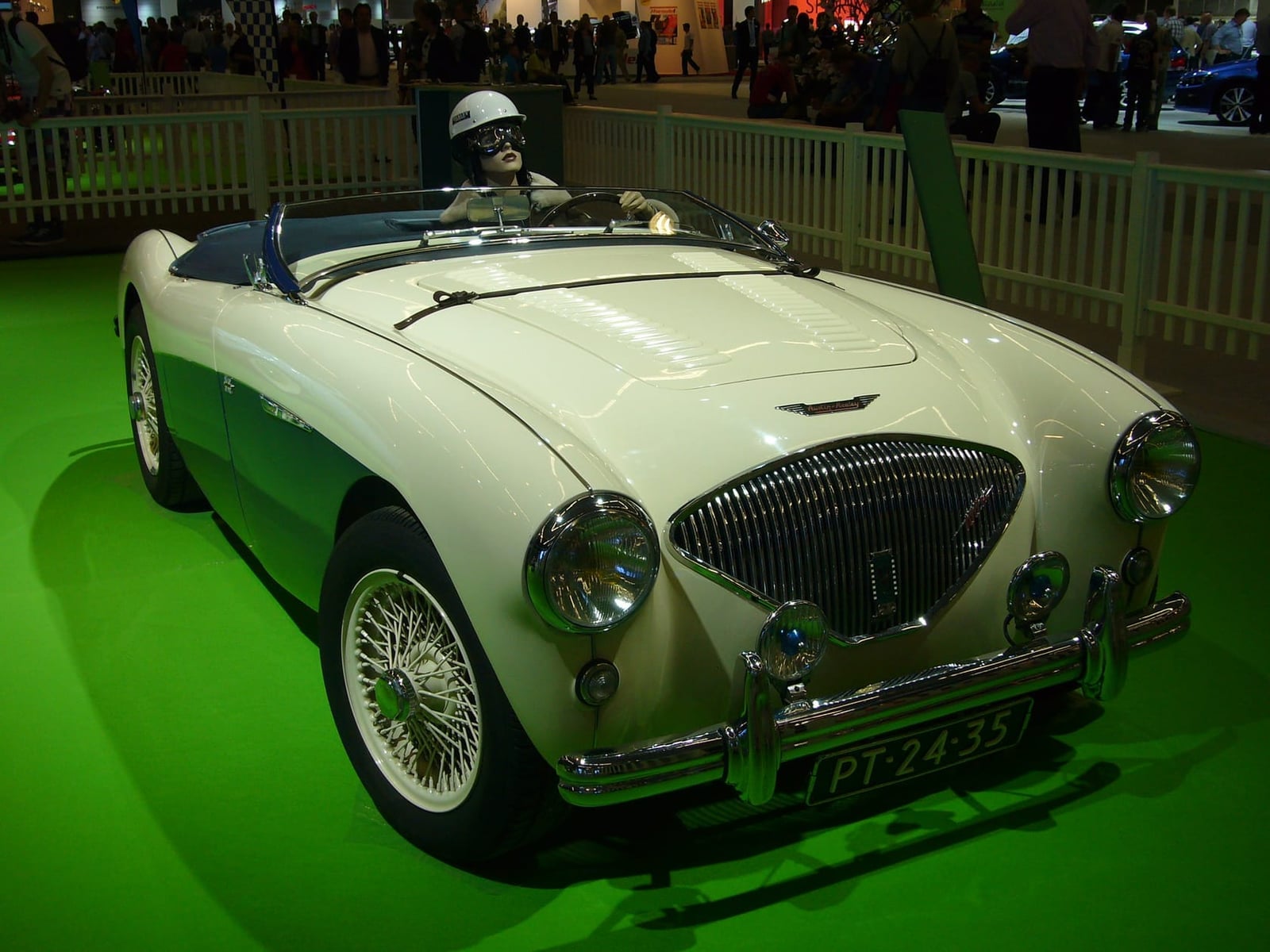 A cream 1954 Austin-Healey 100 on an indoor show stand with a period-dressed mannequin at the wheel