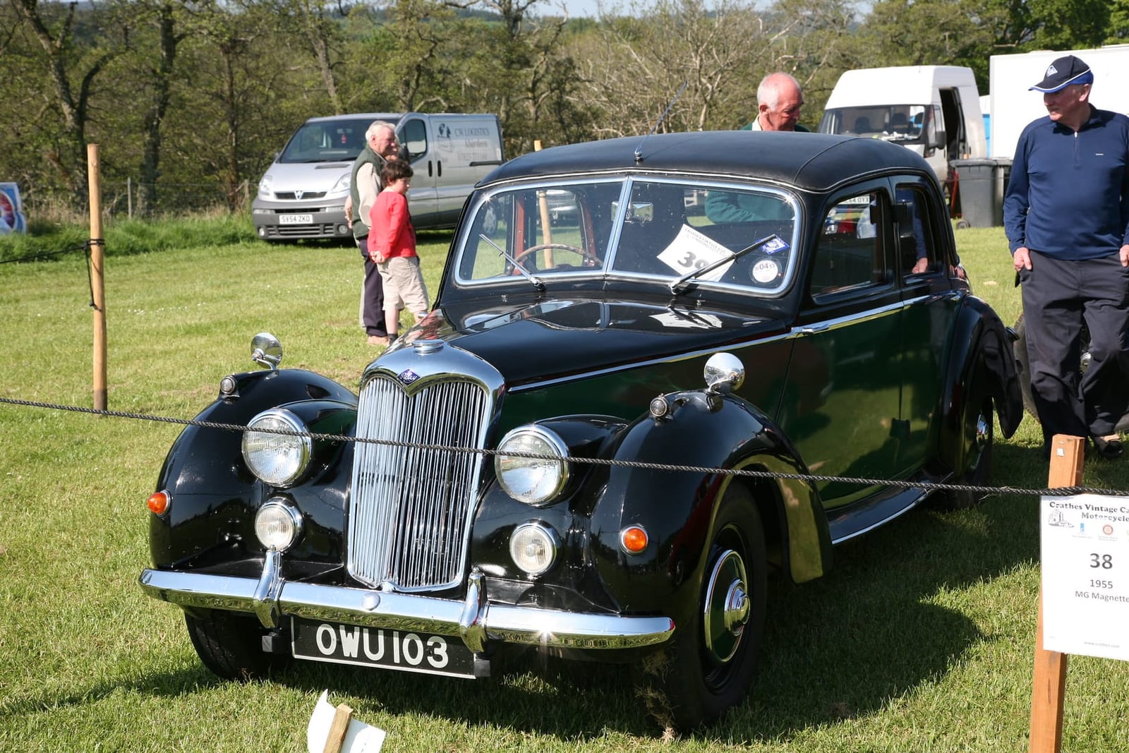 A dark green 1954 Riley RMF saloon on grass at a classic vehicle show, front three-quarter view, UK registration OWU 103