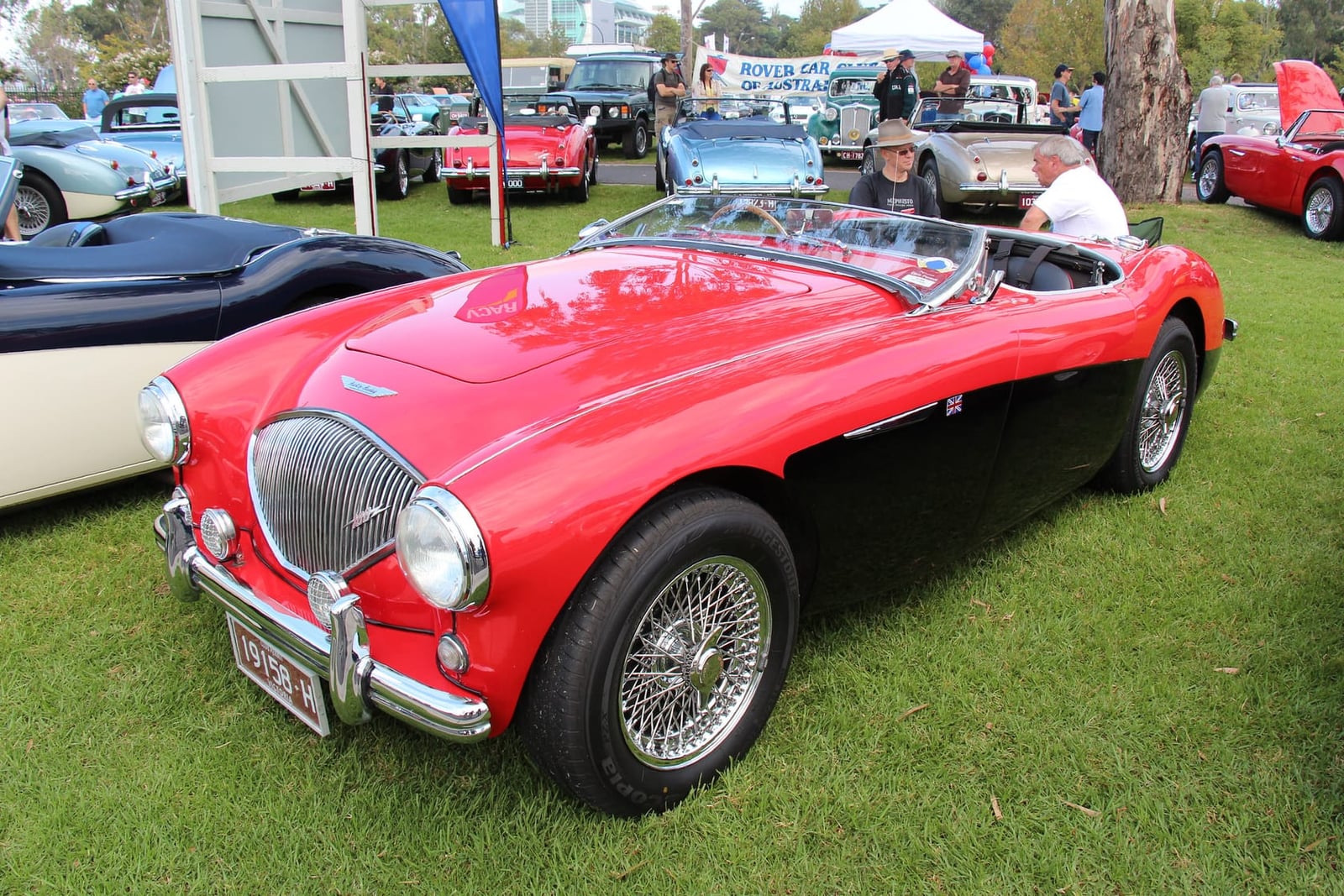 A red 1955 Austin-Healey 100 BN2 roadster on wire wheels at a classic car show, front three-quarter view