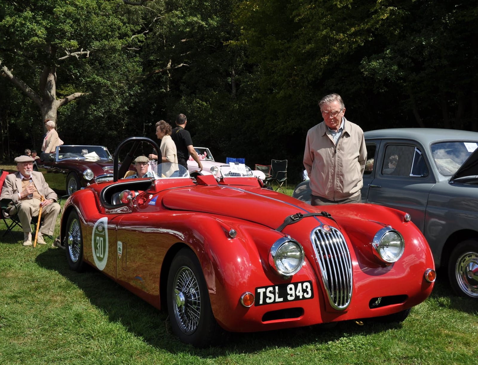 A red 1956 Jaguar XK140 roadster on grass at a classic car show with spectators around it, UK registration TSL 943