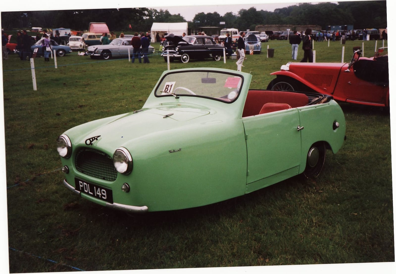 A green 1957 Reliant Regal drophead coupe on grass at a classic vehicle rally, UK registration FDL 149