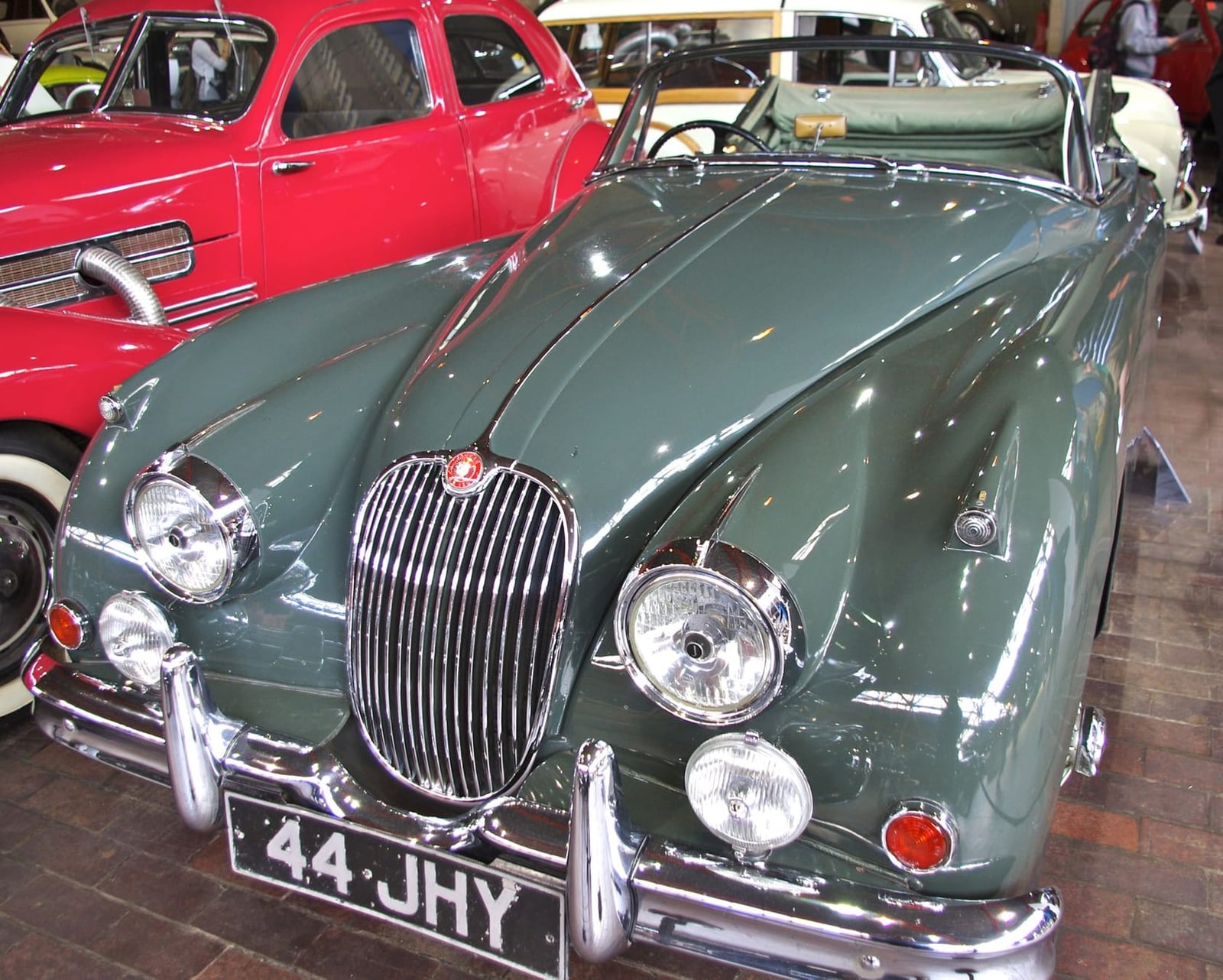 A grey-green Jaguar XK150 roadster on gravel at a classic car event, front three-quarter view, UK registration 44 JHY