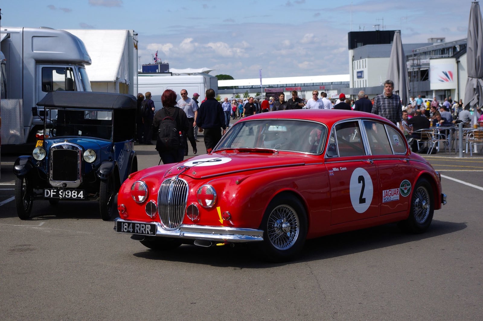 A red Jaguar Mk2 racing saloon, number 2, in a circuit paddock beside a vintage Austin