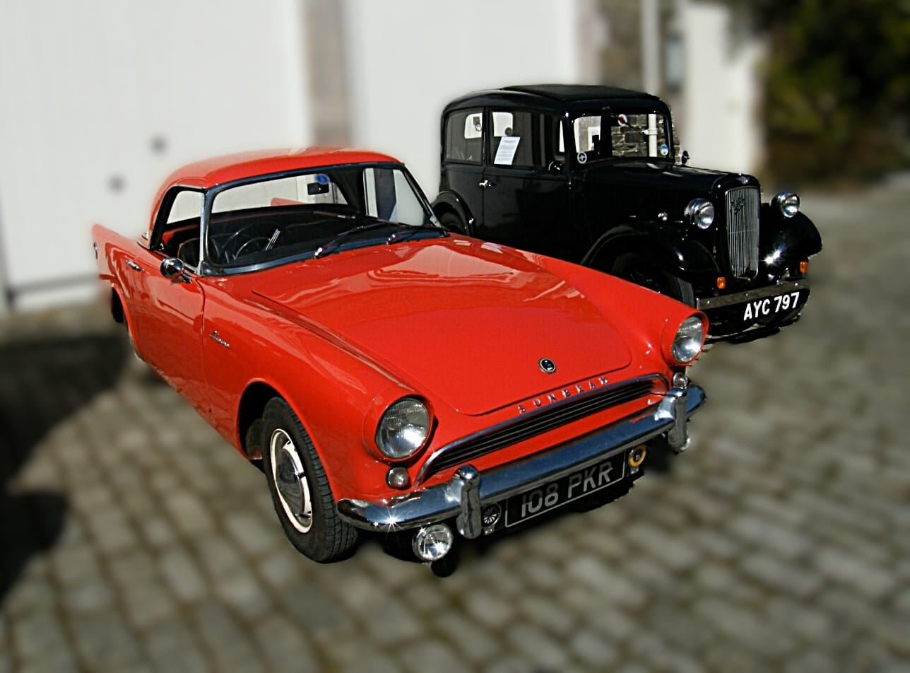 A red 1961 Sunbeam Alpine wearing its black factory hardtop on a cobbled street