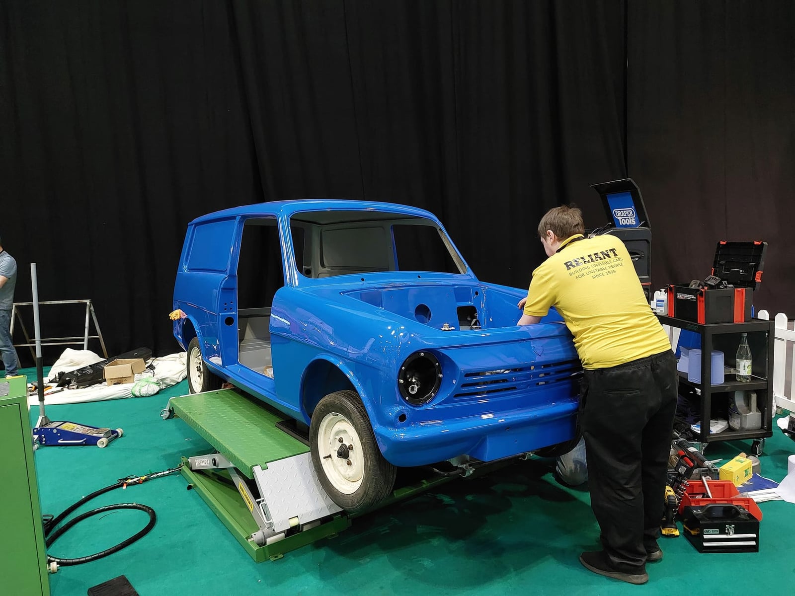 A bare blue Reliant Rebel bodyshell mounted on a rotating stand at a show, with a restorer working on it