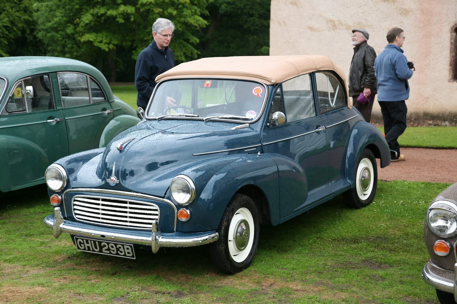 A blue 1964 Morris Minor 1000 convertible with its cream hood raised at a classic car gathering, UK registration GHU 293B