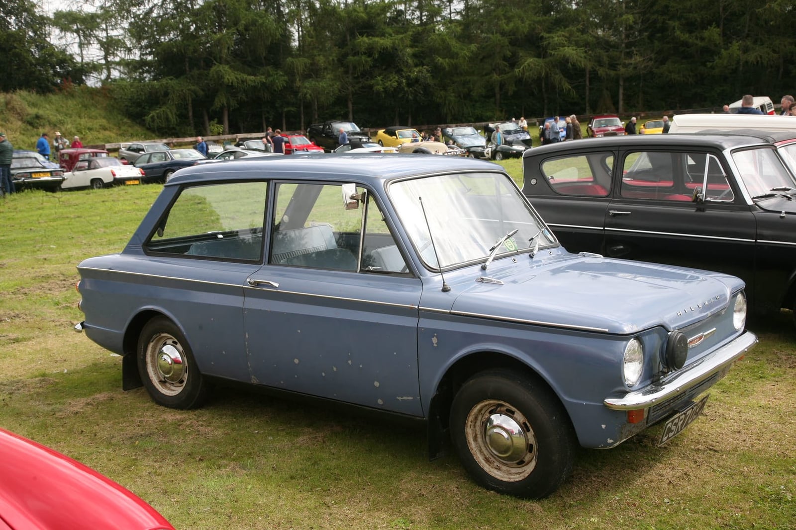An unrestored pale blue 1965 Hillman Imp on grass at a classic gathering, front three-quarter view