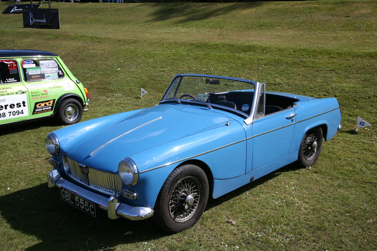 A light blue MG Midget MkII with chrome bumpers and wire wheels, front three-quarter view on grass