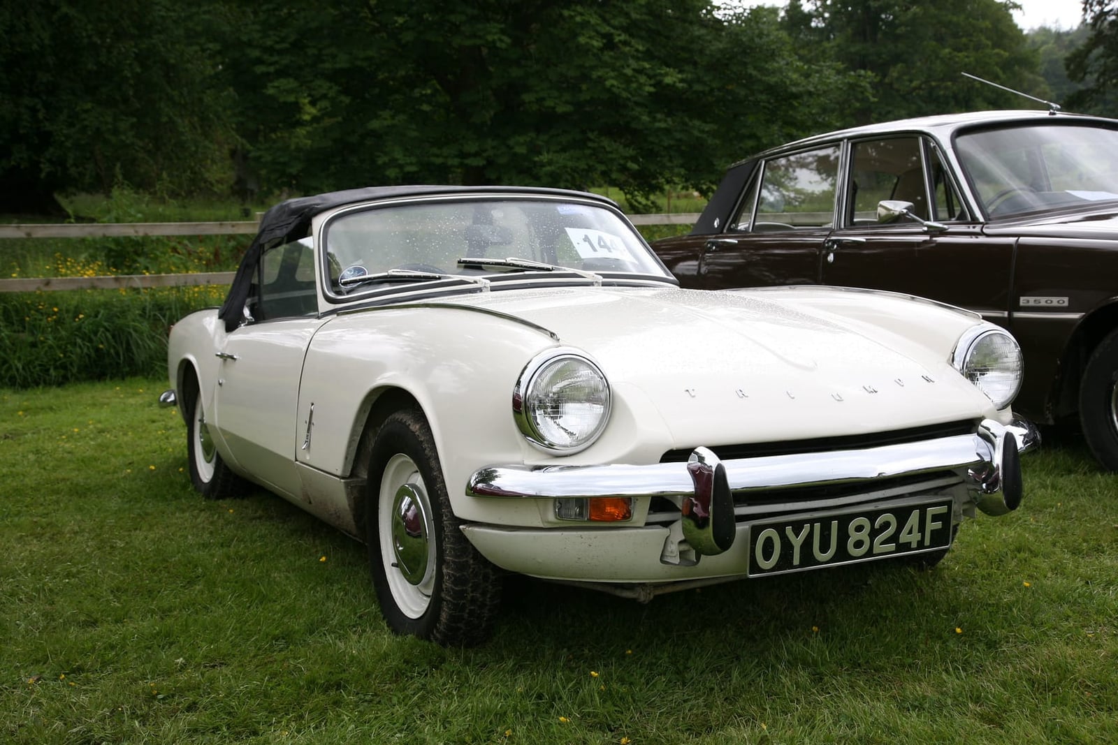 A white 1967 Triumph Spitfire Mk3 with its hood up and a UK registration on a show field
