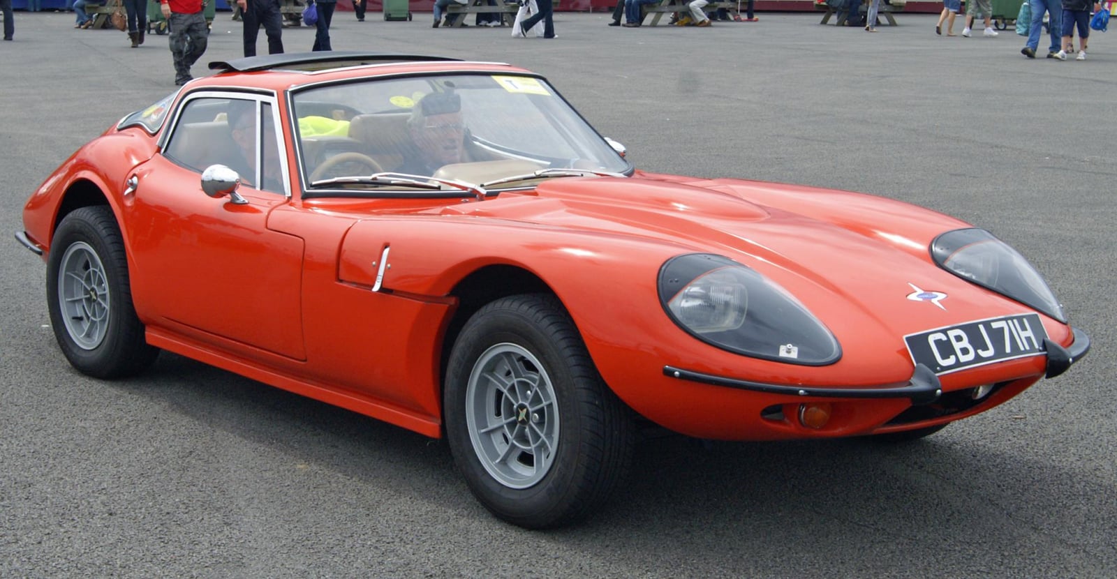 An orange 1969 Marcos GT, front three-quarter view, its low glassfibre fastback body parked at a classic-car show