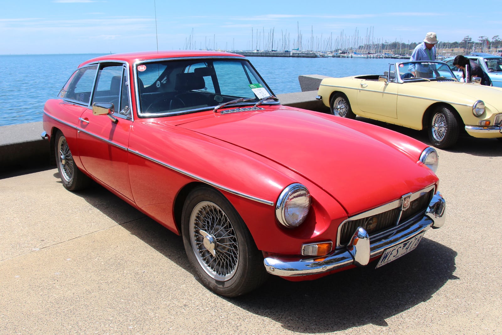 A red early MG MGB GT, front three-quarter view by a harbour