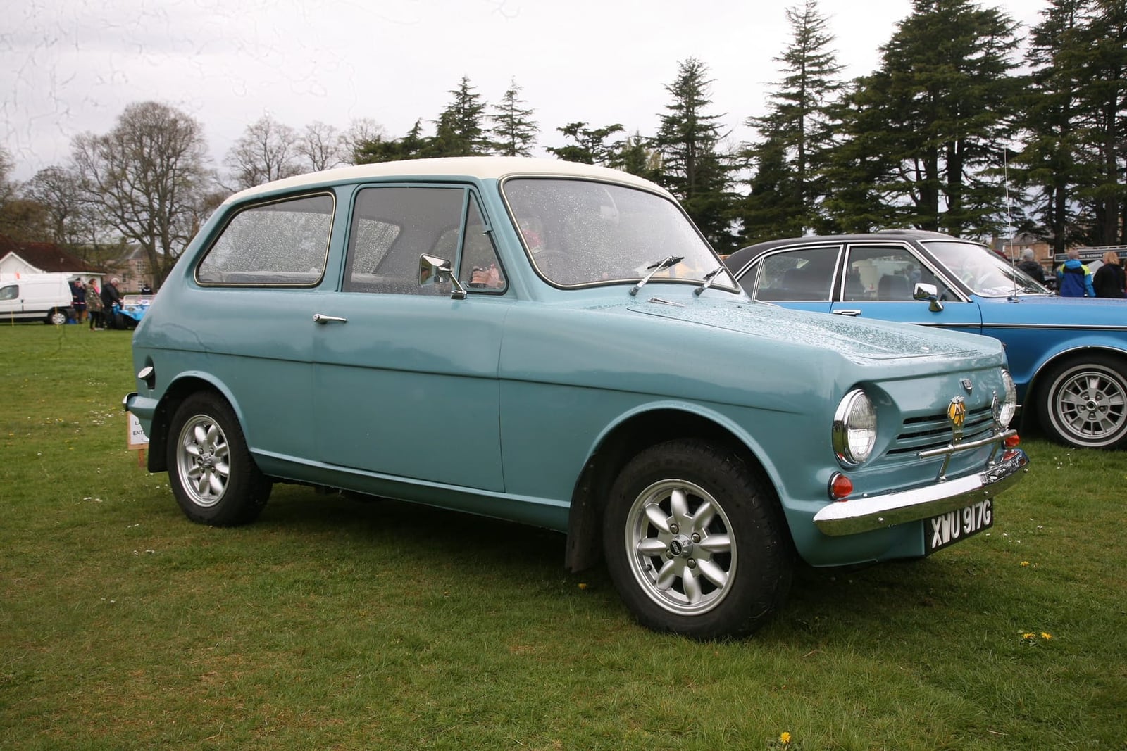 A light blue Reliant Rebel 700 estate on grass at a classic vehicle show, front three-quarter view, UK registration XWU 917G