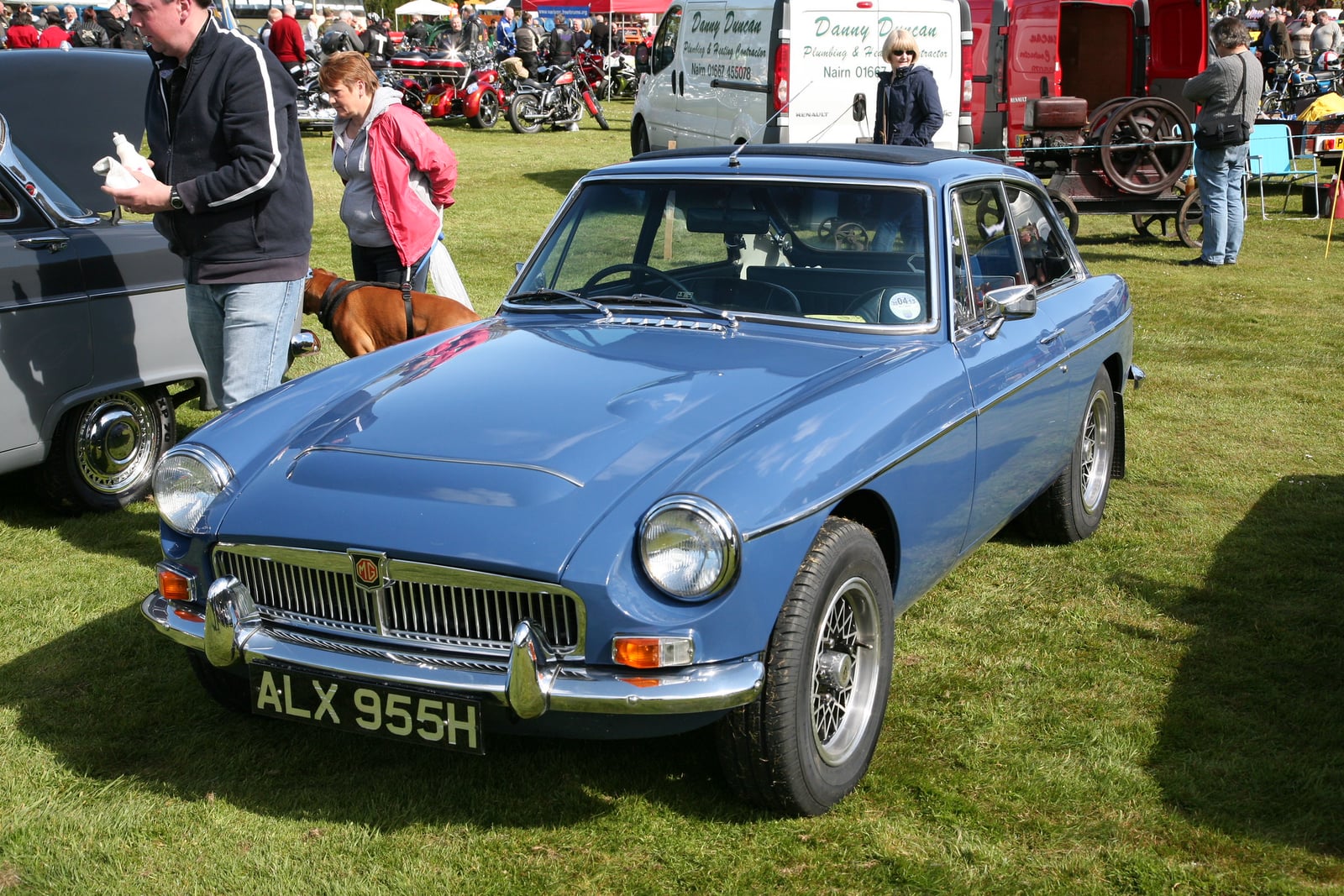 A light blue MG MGC GT, front three-quarter view on grass, showing the bonnet bulge