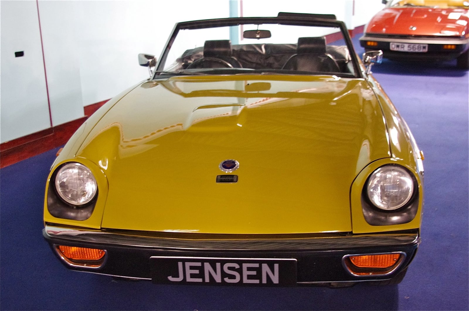 A yellow Jensen-Healey at a show in front of a Jensen banner, front three-quarter view
