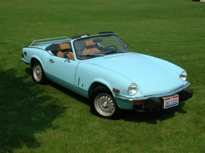 A pale blue Triumph Spitfire 1500 with a luggage rack and tan interior, top down, front three-quarter view on grass