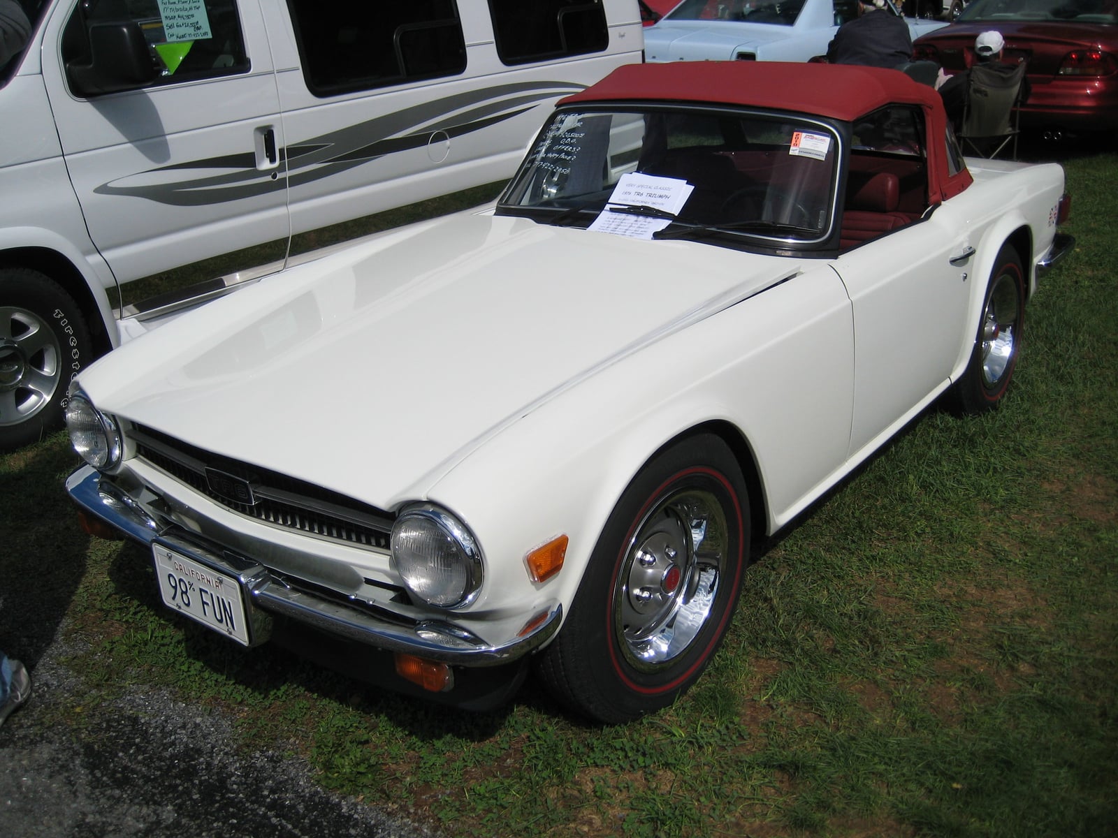 A white Triumph TR6 with a red soft top, front three-quarter view on grass at a show