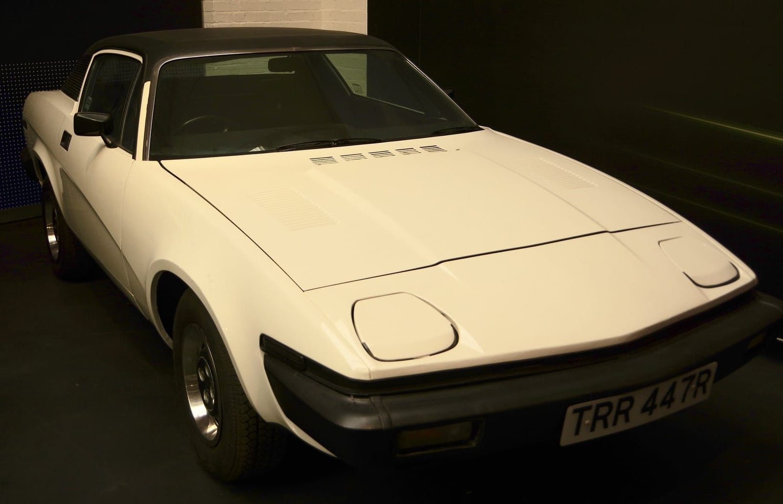 A white 1976 Triumph TR7 fixed-head coupe with a UK registration, photographed indoors against a dark wall