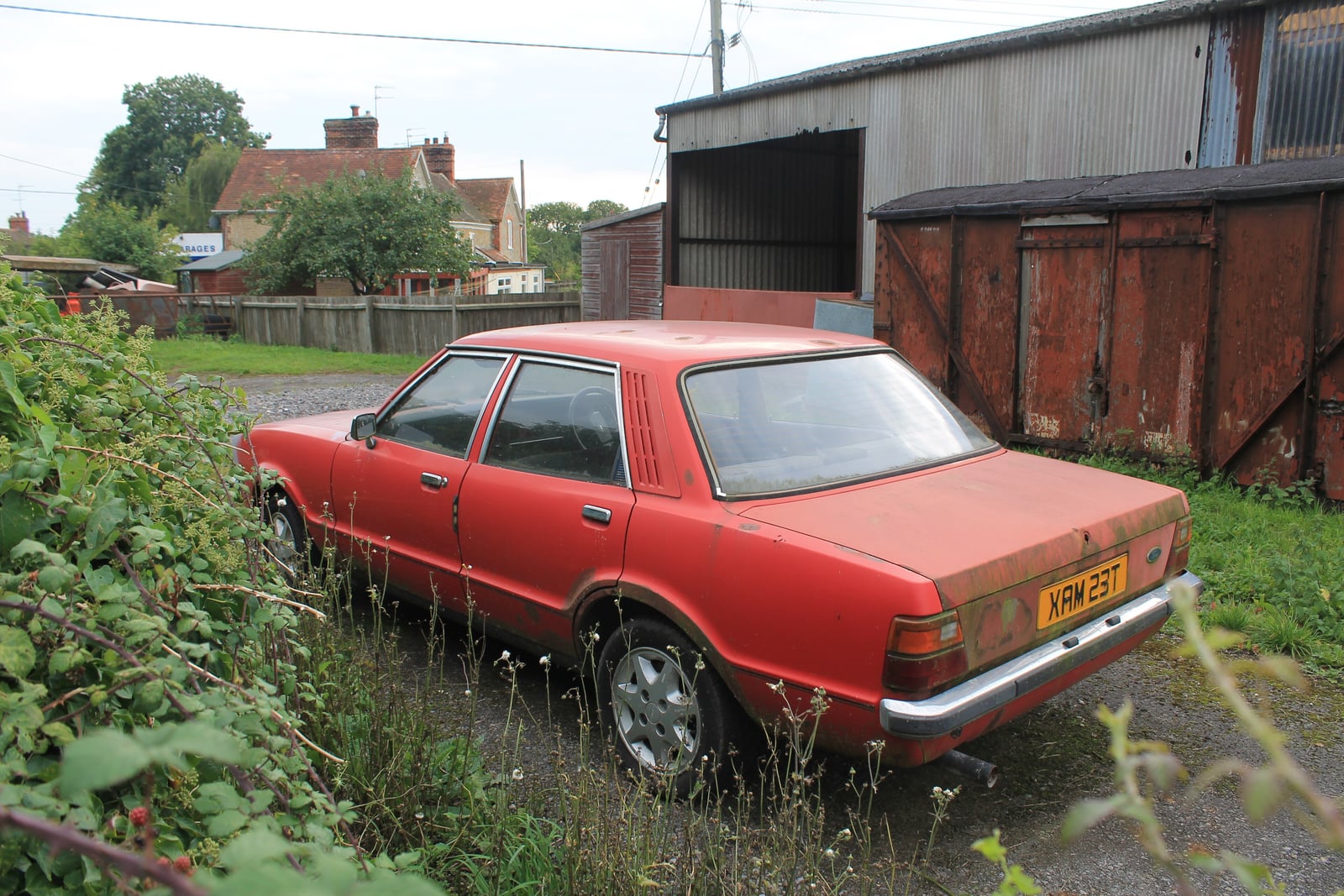 A faded red Ford Cortina Mk4 awaiting restoration in a yard, rear three-quarter view