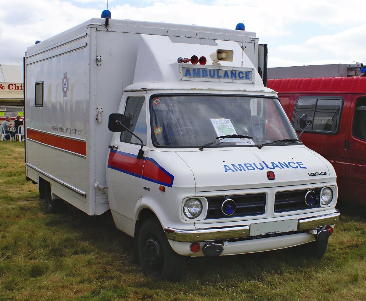 A white 1980 Bedford CF ambulance in Dorset Ambulance Service livery with a blue roof beacon bar, front three-quarter view on grass at a show