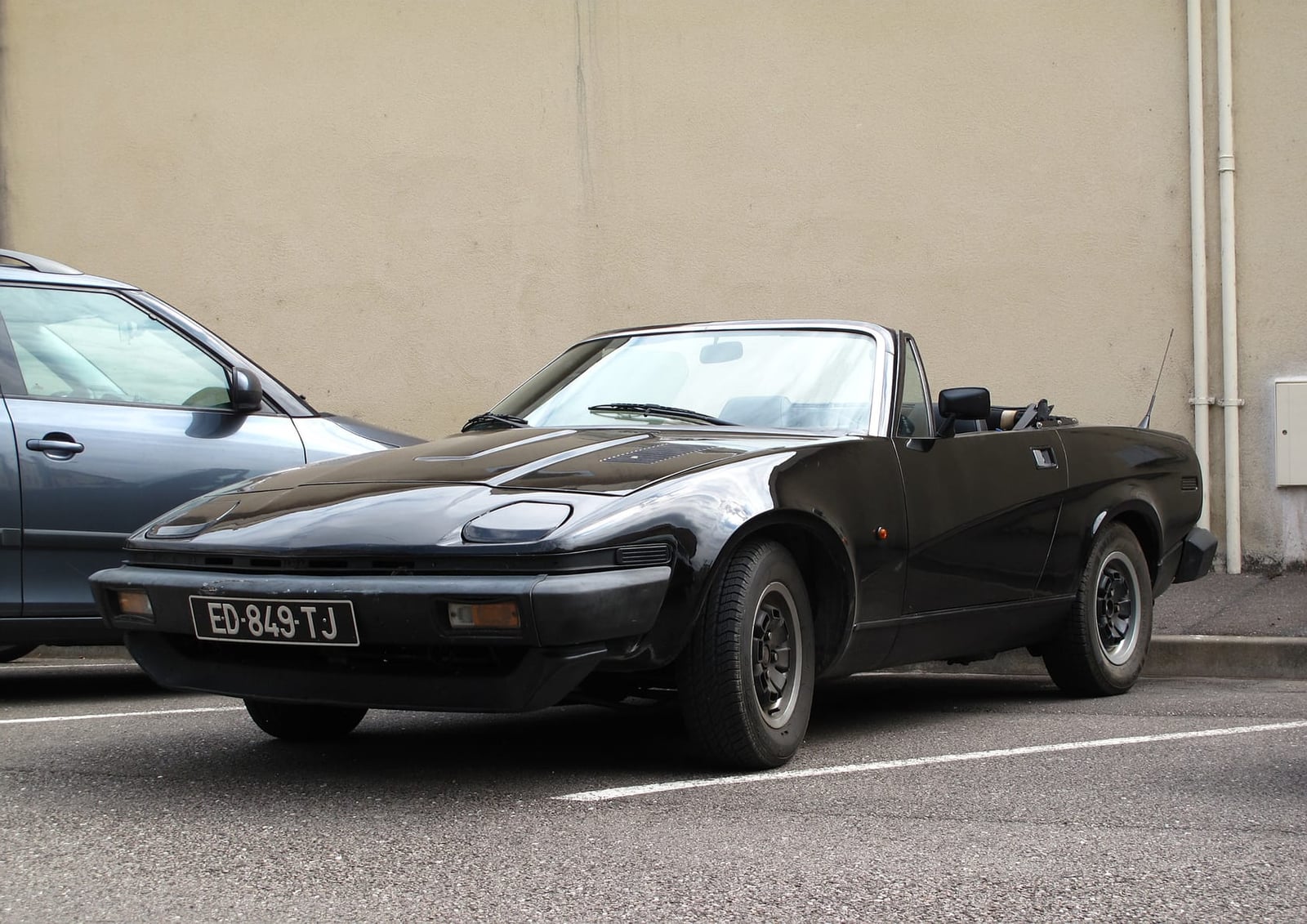 A black Triumph TR7 convertible with a UK registration parked against a plain wall