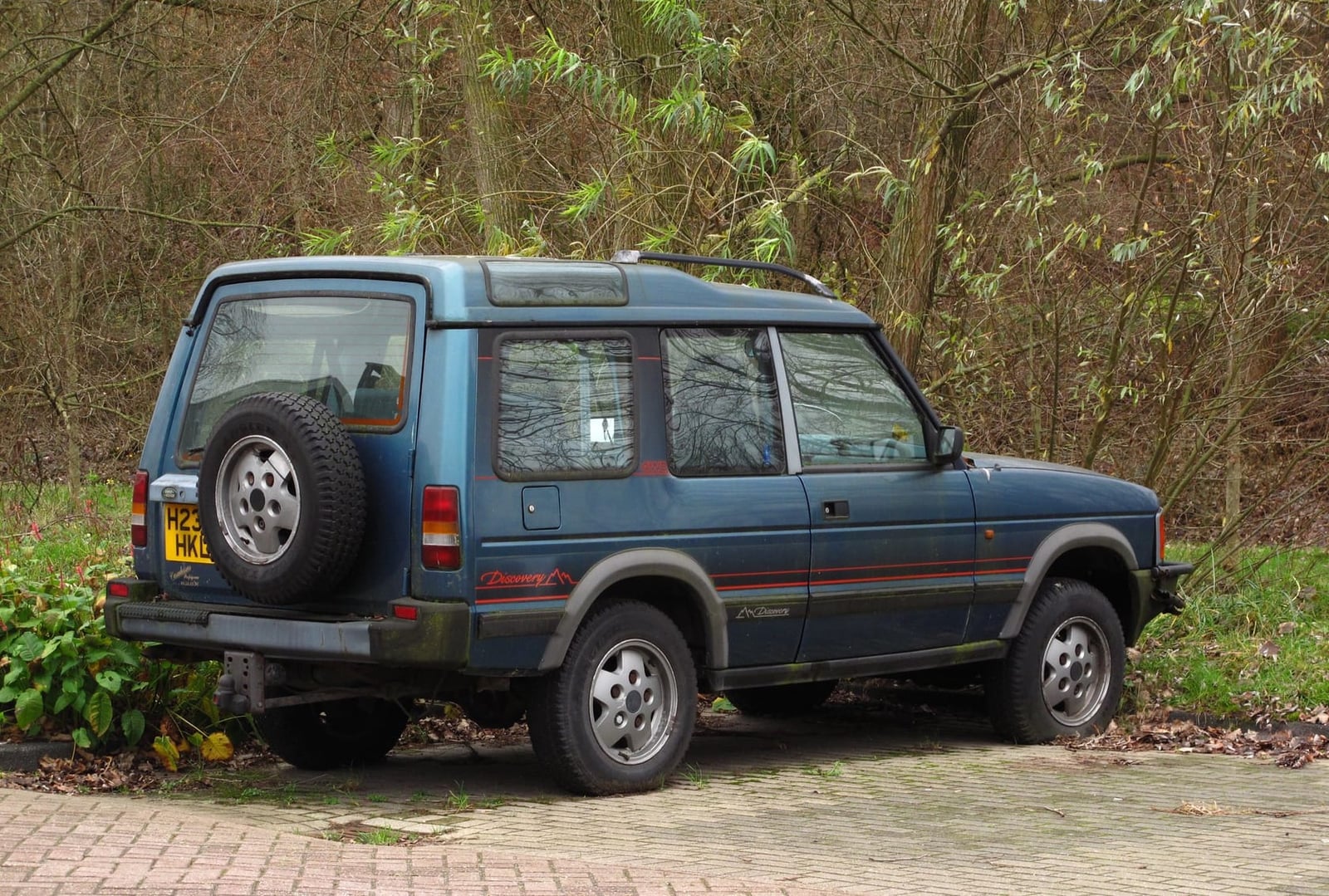 An early 1990 Land Rover Discovery parked under trees, rear three-quarter view with spare wheel on the tailgate