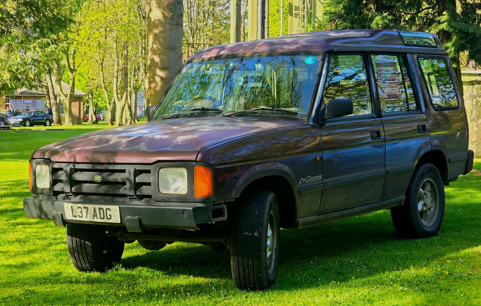 A maroon 1994 Land Rover Discovery V8i with sun-faded paint parked on grass under trees