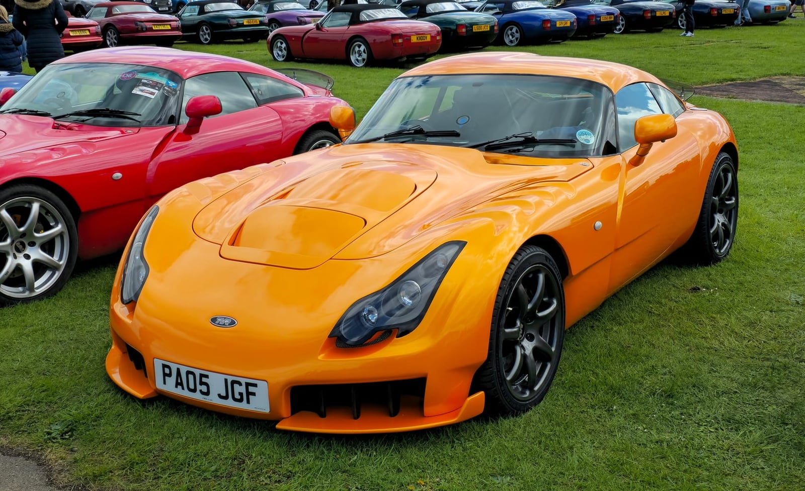 An orange 2005 TVR Sagaris with a UK registration at a gathering of TVRs on grass, with a row of Blackpool cars behind