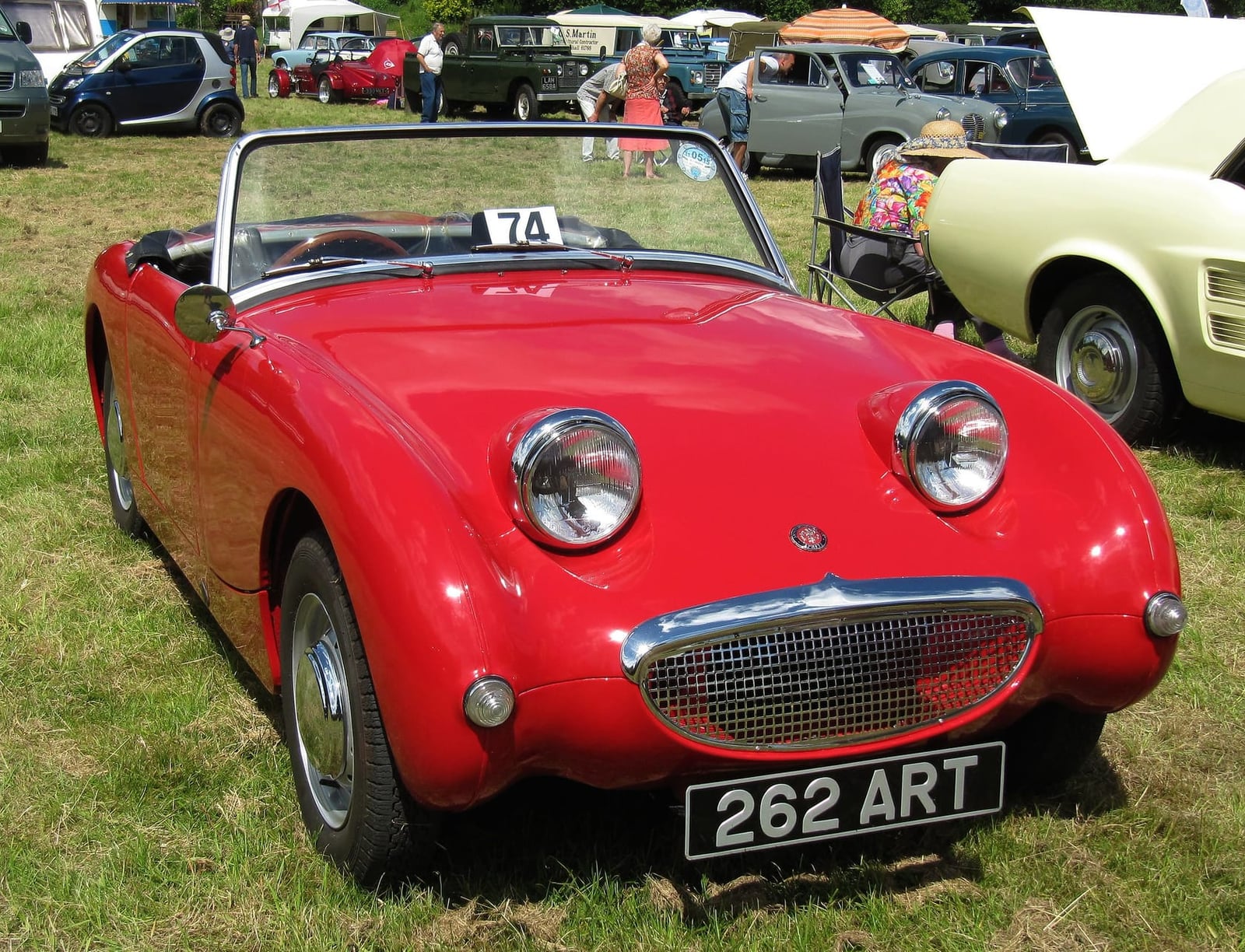 A red Austin-Healey Frogeye Sprite on grass at a classic vehicle rally, front three-quarter view, UK registration 262 ART