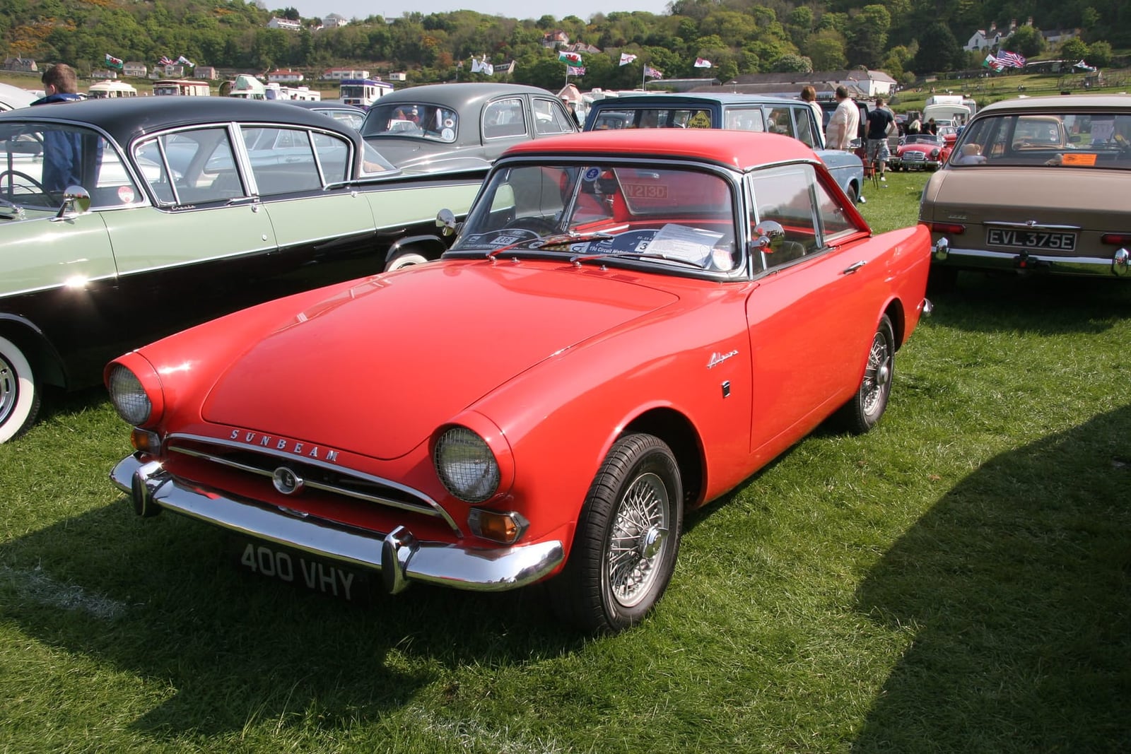A red 1964 Sunbeam Alpine on wire wheels at a classic car show, front three-quarter view, UK registration 400 VHY
