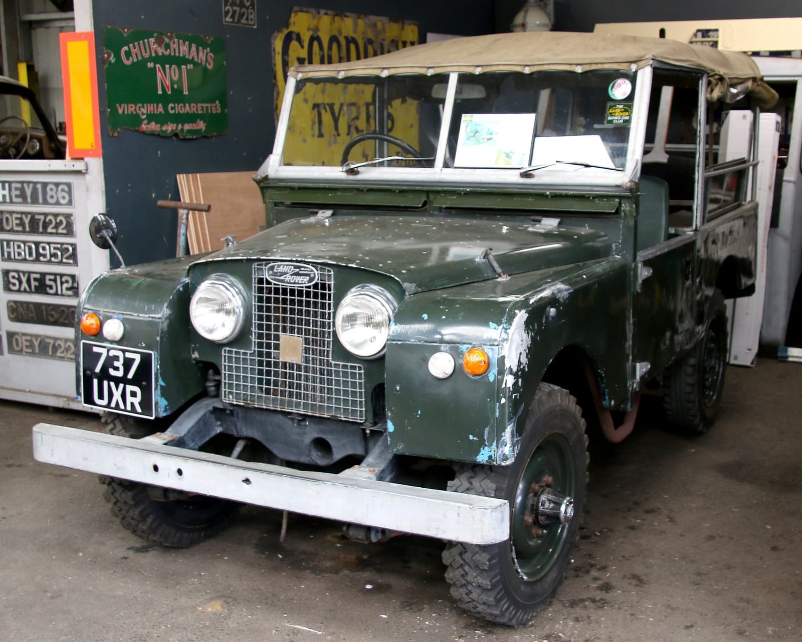A patinated green 1957 Land Rover Series I awaiting restoration inside a workshop with period signs