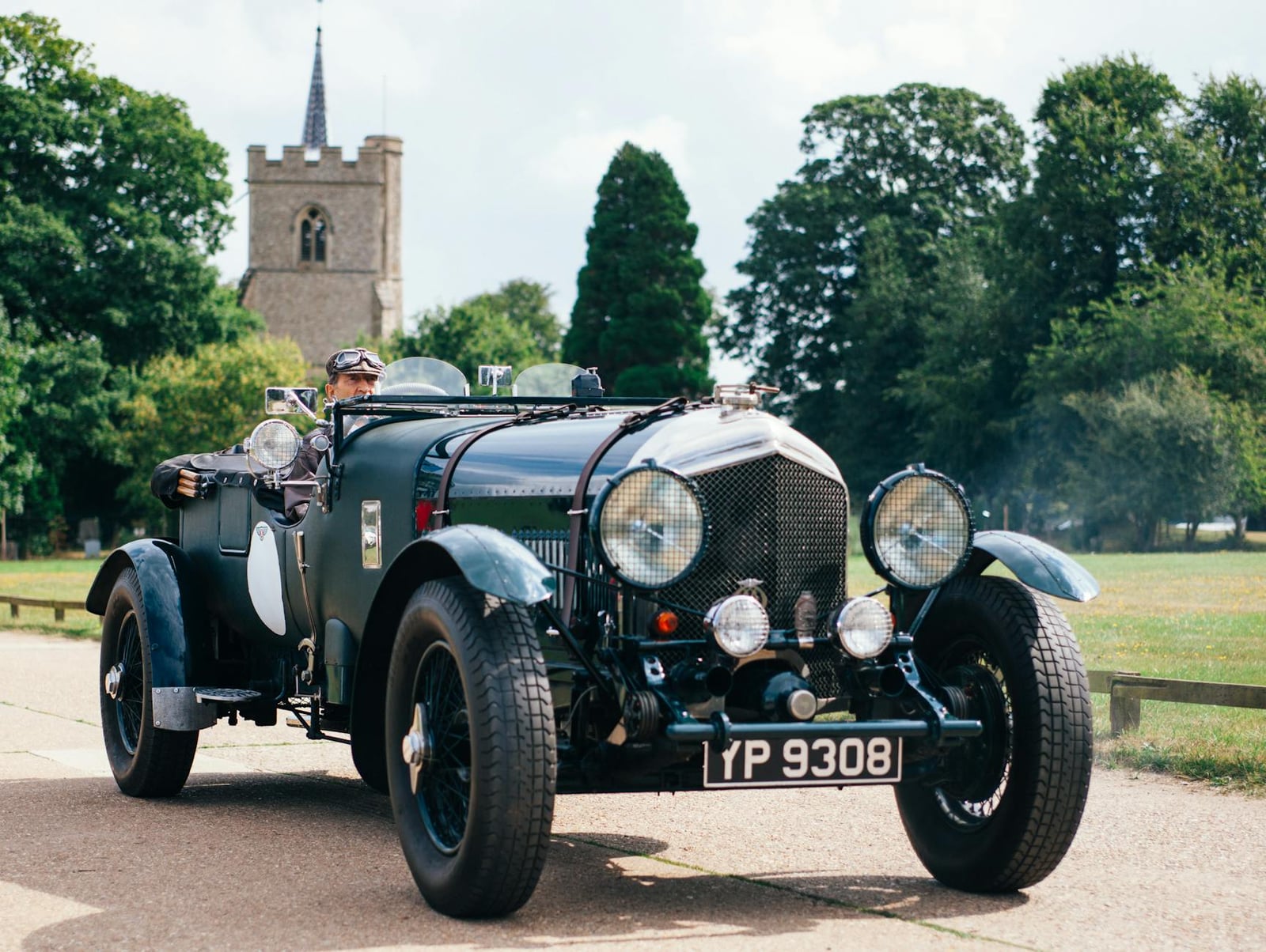A green pre-war Bentley open tourer with a goggled driver, driving past a village church, registration "YP 9308"