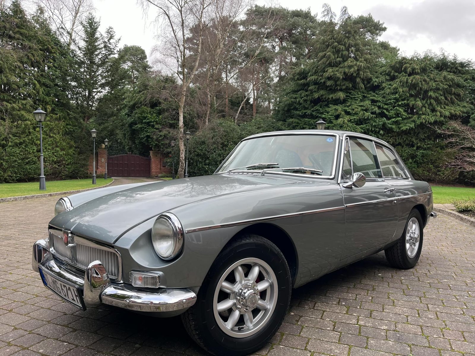 A silver MGB GT coupe with chrome bumpers and Rostyle wheels, parked on a driveway with woodland behind