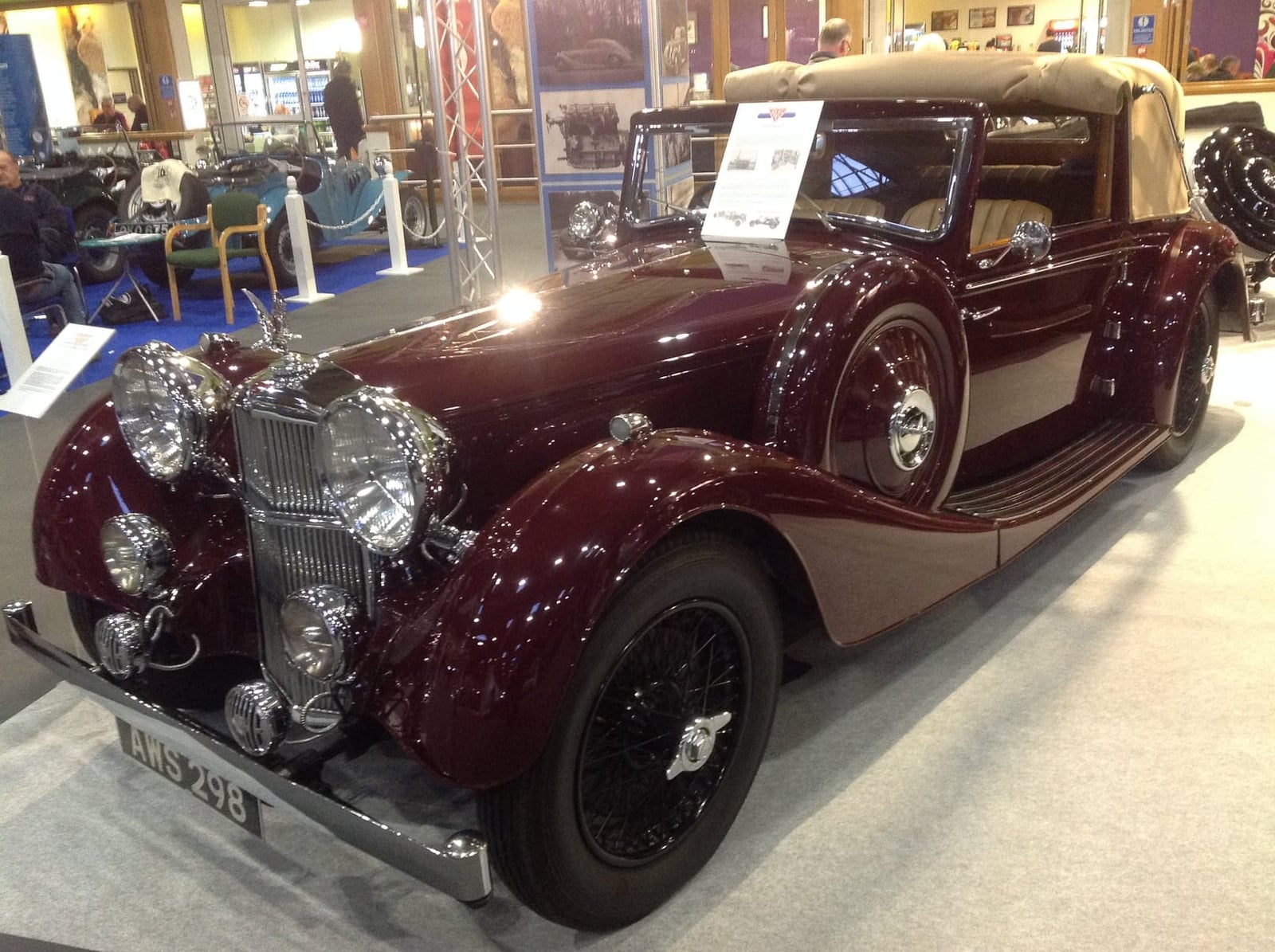 A maroon 1937 Alvis Speed 25 drophead coupe with its cream hood raised, on display indoors at a classic car show, front three-quarter view, UK registration AWS 298