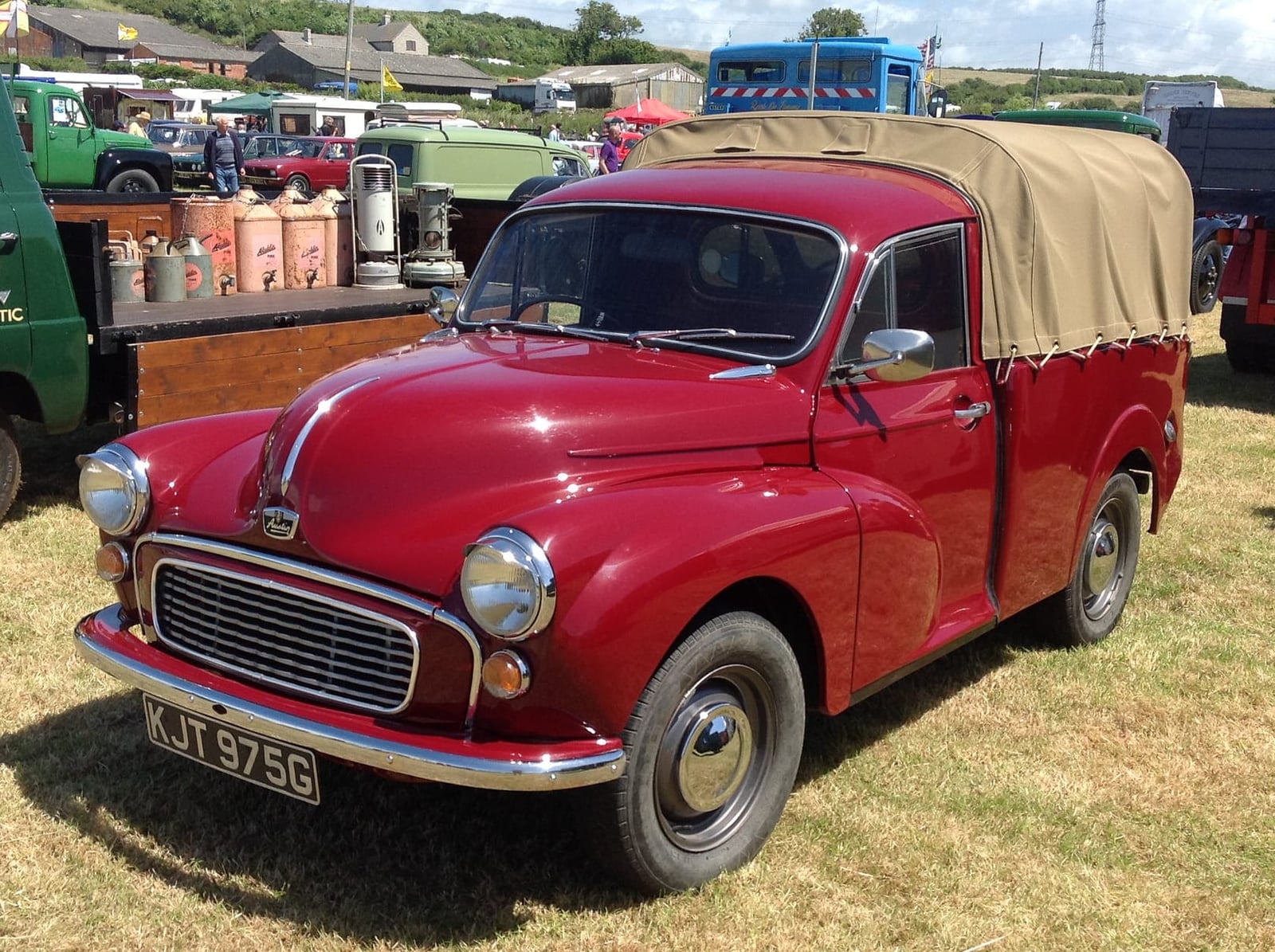 A maroon 1969 Minor-based 6 cwt pickup with a canvas tilt over the bed, on grass at a commercial vehicle rally, UK registration KJT 975G