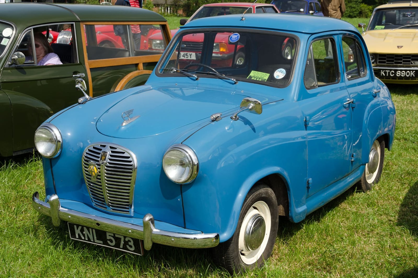 A blue 1956 Austin A30 four-door saloon parked on grass at a classic car show, front three-quarter view, UK registration KNL 573