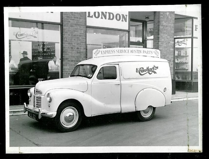 A period black and white photograph of a white Austin A40 van parked outside an Austin parts shop in London