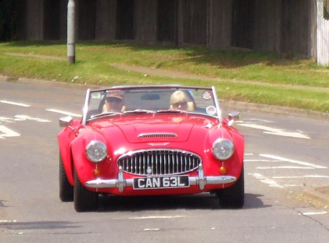 A red Austin-Healey 100 being driven on a road with two occupants aboard, front view, UK registration CAN 63L