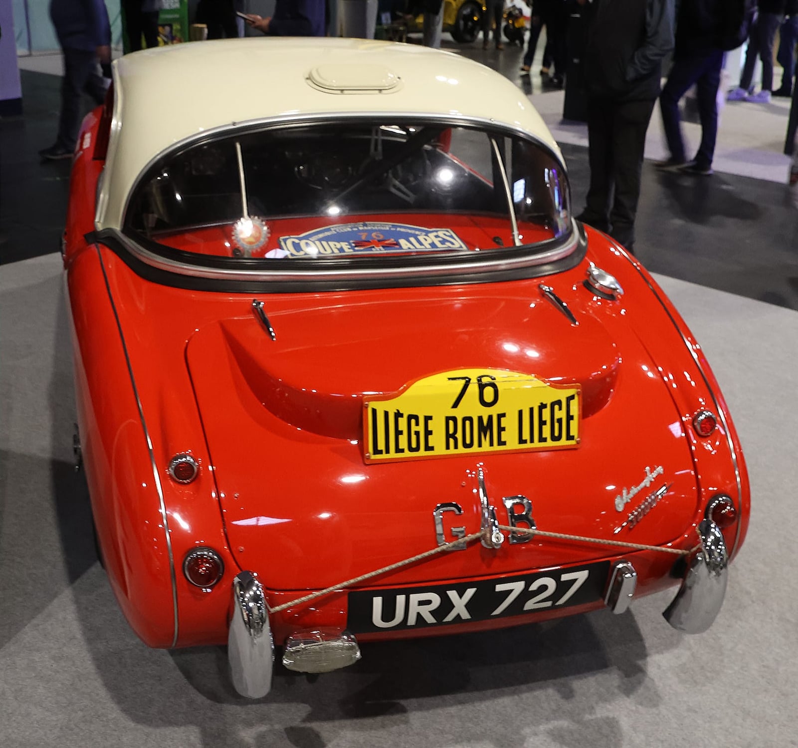 The rear of a red 1960 Austin-Healey 3000 works rally car wearing its Liège-Rome-Liège rally plate, displayed at an indoor show