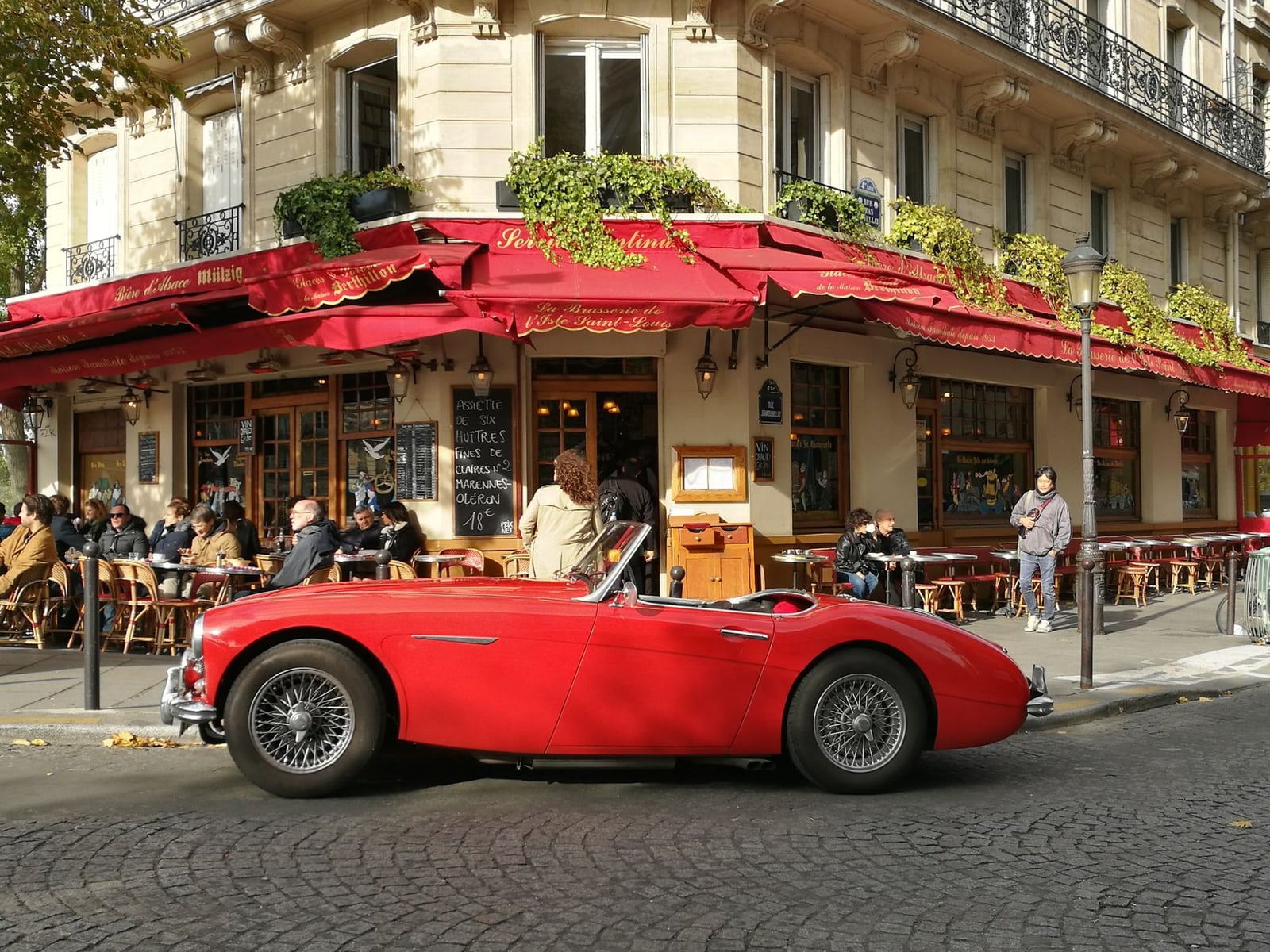 A red Austin-Healey roadster with wire wheels and the top folded down, parked at the kerb outside a busy cafe