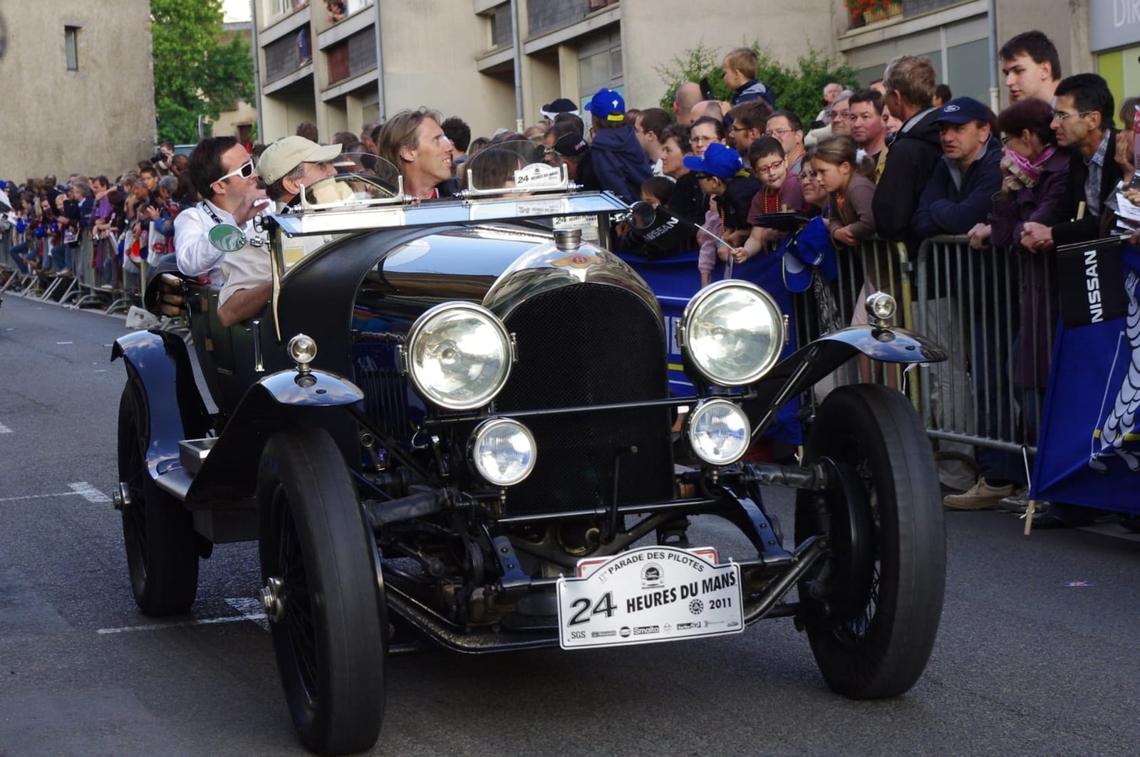 A dark green Blower Bentley leading a parade at the Le Mans Classic with crowds behind
