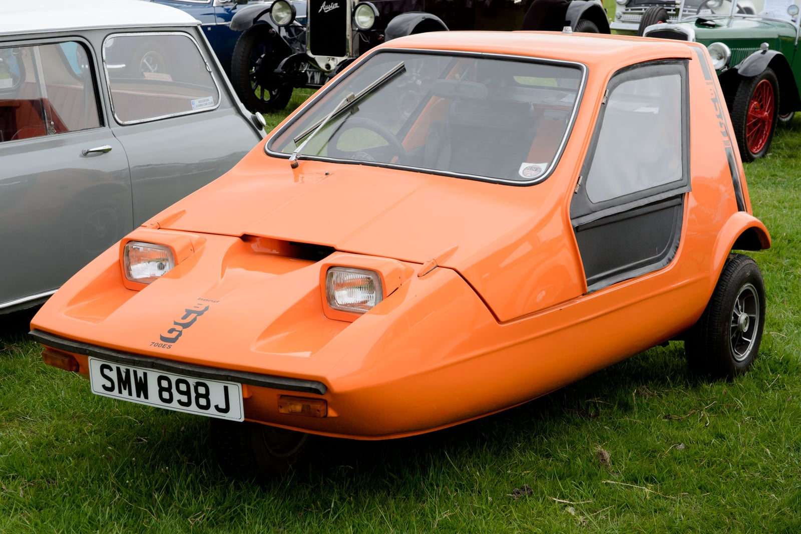 An orange 1970 Bond Bug (SMW898J) on grass beside a classic Mini, front three-quarter view