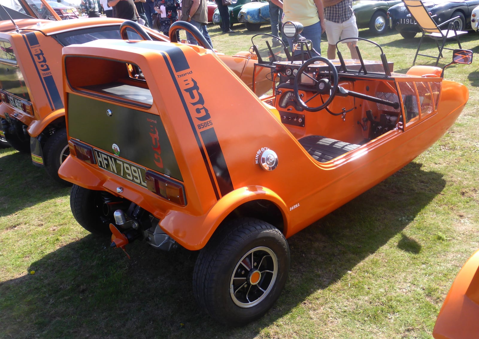 An orange 1973 Bond Bug (AFN799L) from the rear three-quarter on grass at a show, the open cockpit and side stripe visible