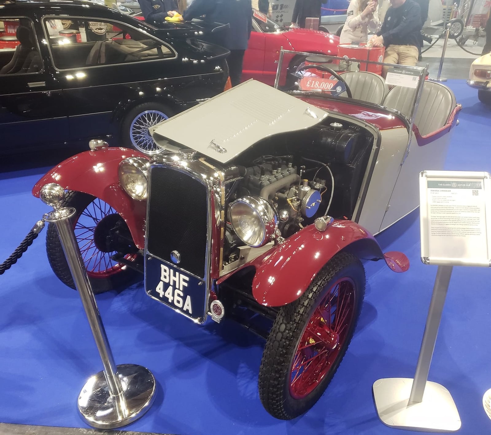 A maroon 1934 BSA three-wheeler on indoor display with its bonnet side removed, showing the v-twin engine ahead of the front axle, UK registration BHF 446A
