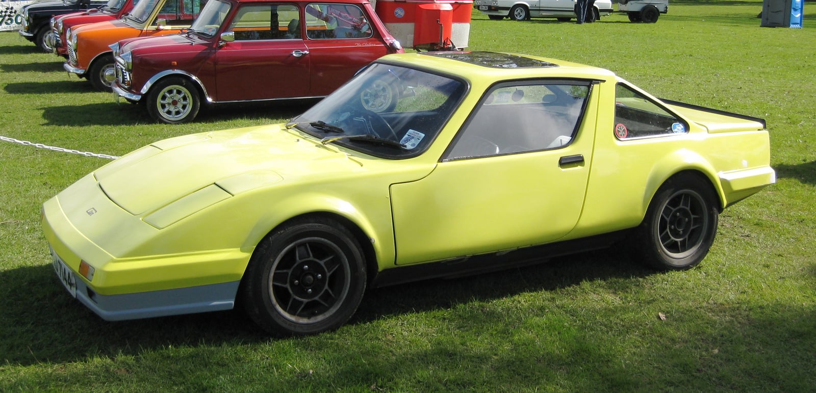 A bright yellow Clan Crusader coupe, front three-quarter view on grass at a classic car show with Minis behind