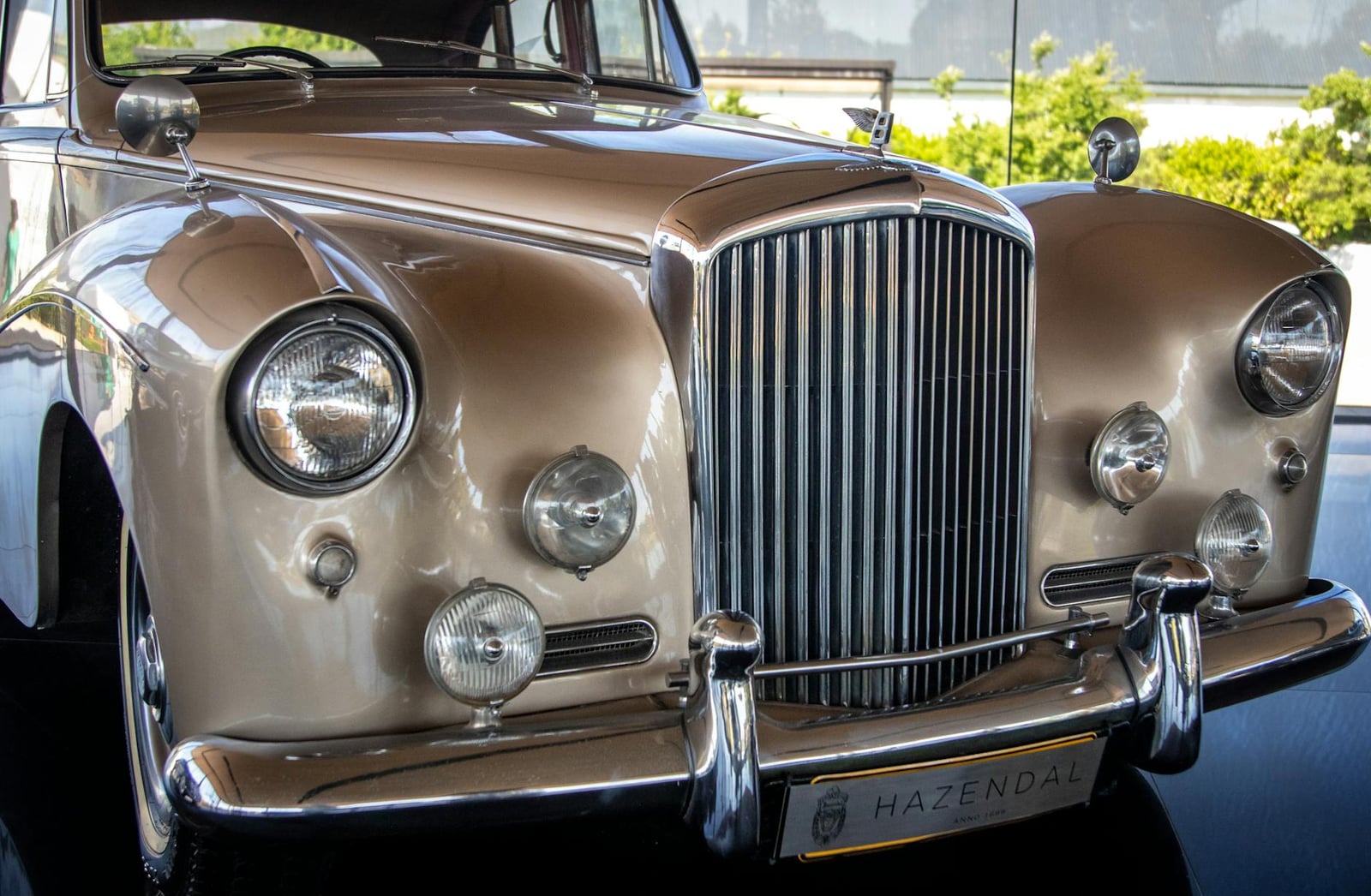 The front of a champagne-gold Bentley saloon, chrome grille and round headlamps, in a showroom