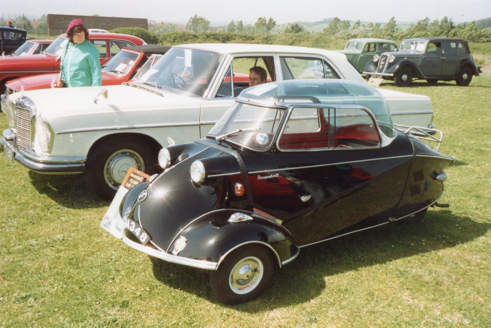 A black and green Messerschmitt Kabinenroller parked on grass beside a white Mercedes-Benz saloon